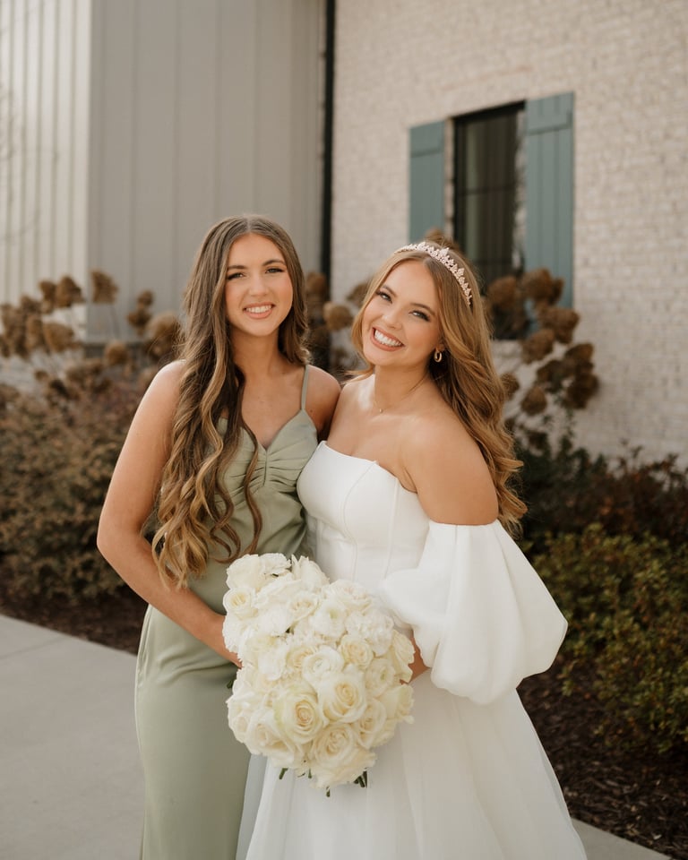 Two smiling women posing together outdoors, one wearing a white wedding dress with tiara holding a white rose bouquet, the other in a sage green dress
