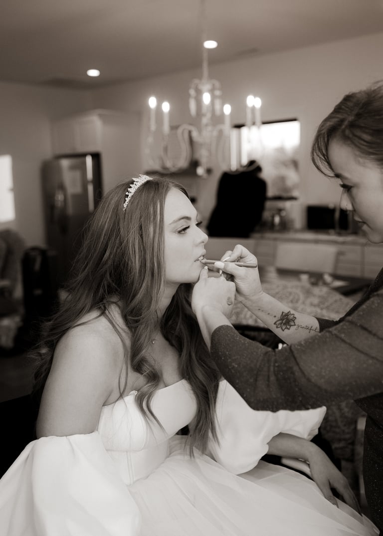 Makeup artist applies makeup to bride in white dress wearing pearl headpiece in bridal suite with candles
