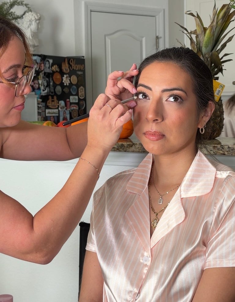 A makeup artist applies eyeshadow to a woman wearing a pink blouse in a bright room with plants and a photo board in the background.