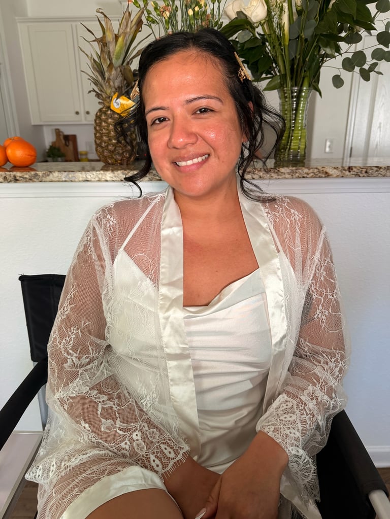 Bridal transformation before - woman smiling at camera wearing white blouse and lace cardigan, seated indoors with kitchen and flowers in background