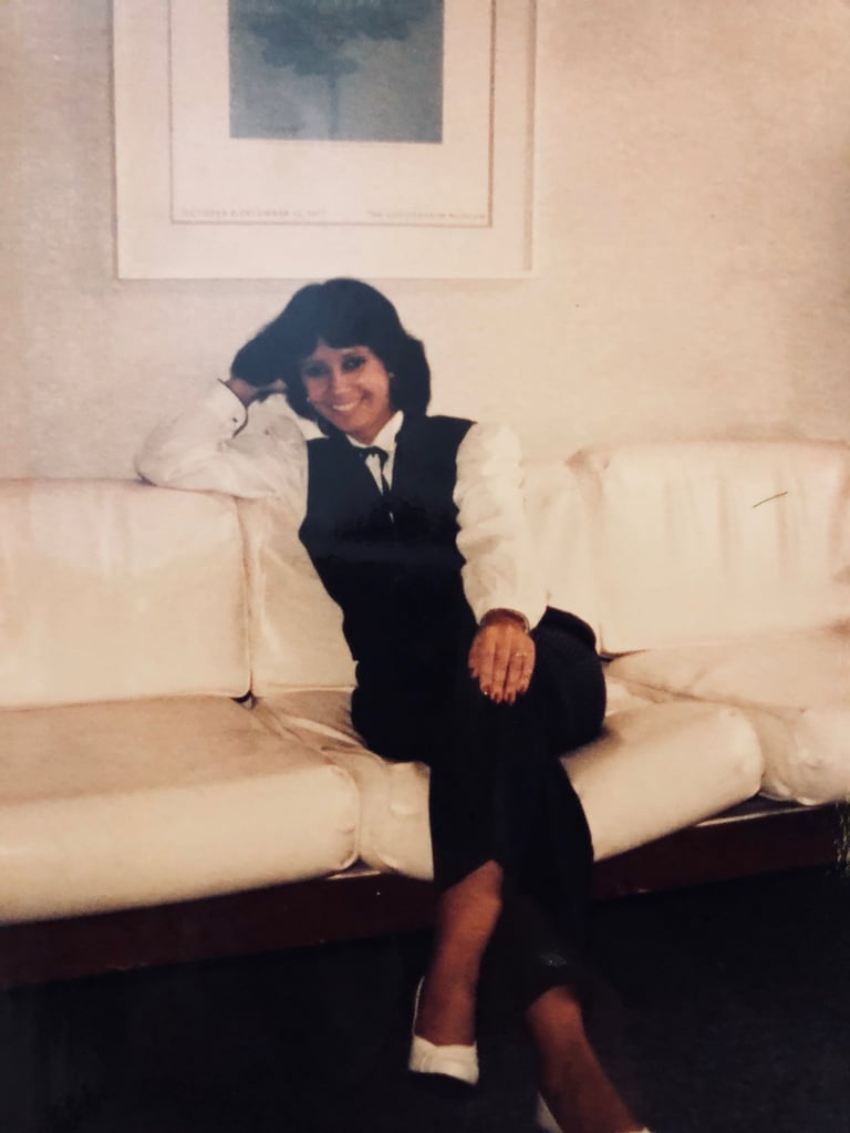 Woman with dark hair wearing a black vest and white blouse sitting on a cream-colored sofa, smiling at the camera
