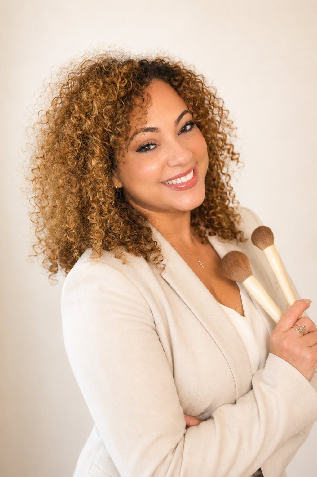 Suzette, makeup artist with curly auburn hair, wearing cream blazer and smiling warmly at camera
